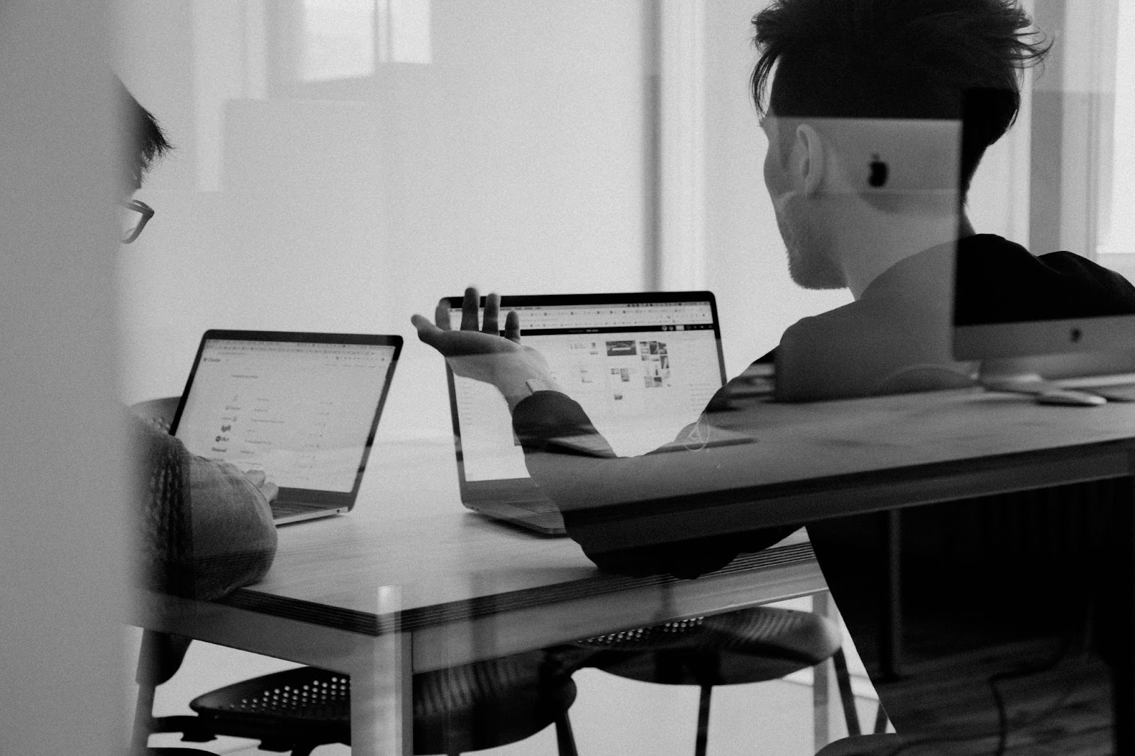 Two men sit at a table with their desktop computers. One man has his arm raised in exasperation.