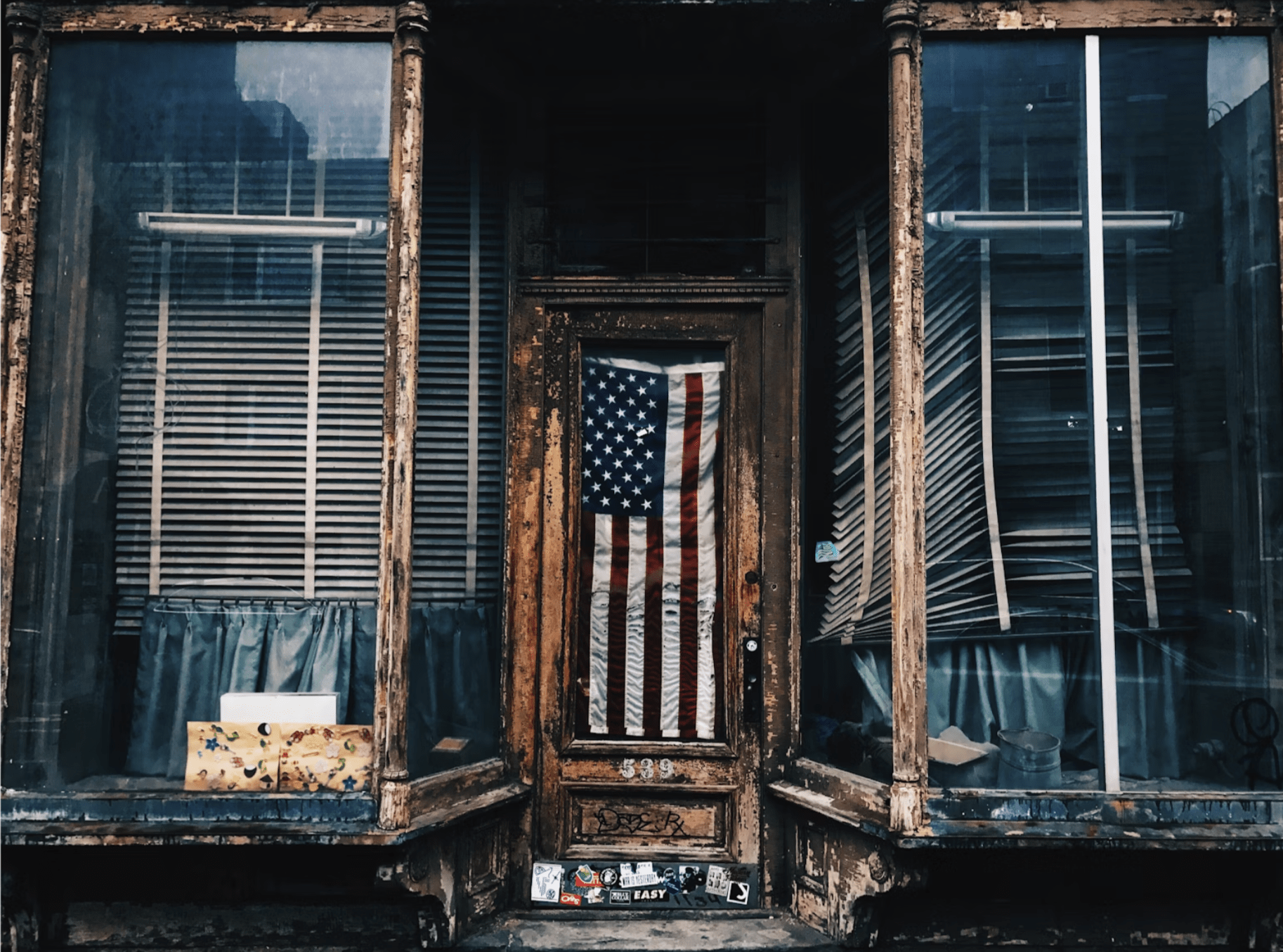A deserted and run-down shopfront has a well kept American flag hanging in the door. Paint is peeling, wood is rotting, and the window shades are barely functional on one display case.