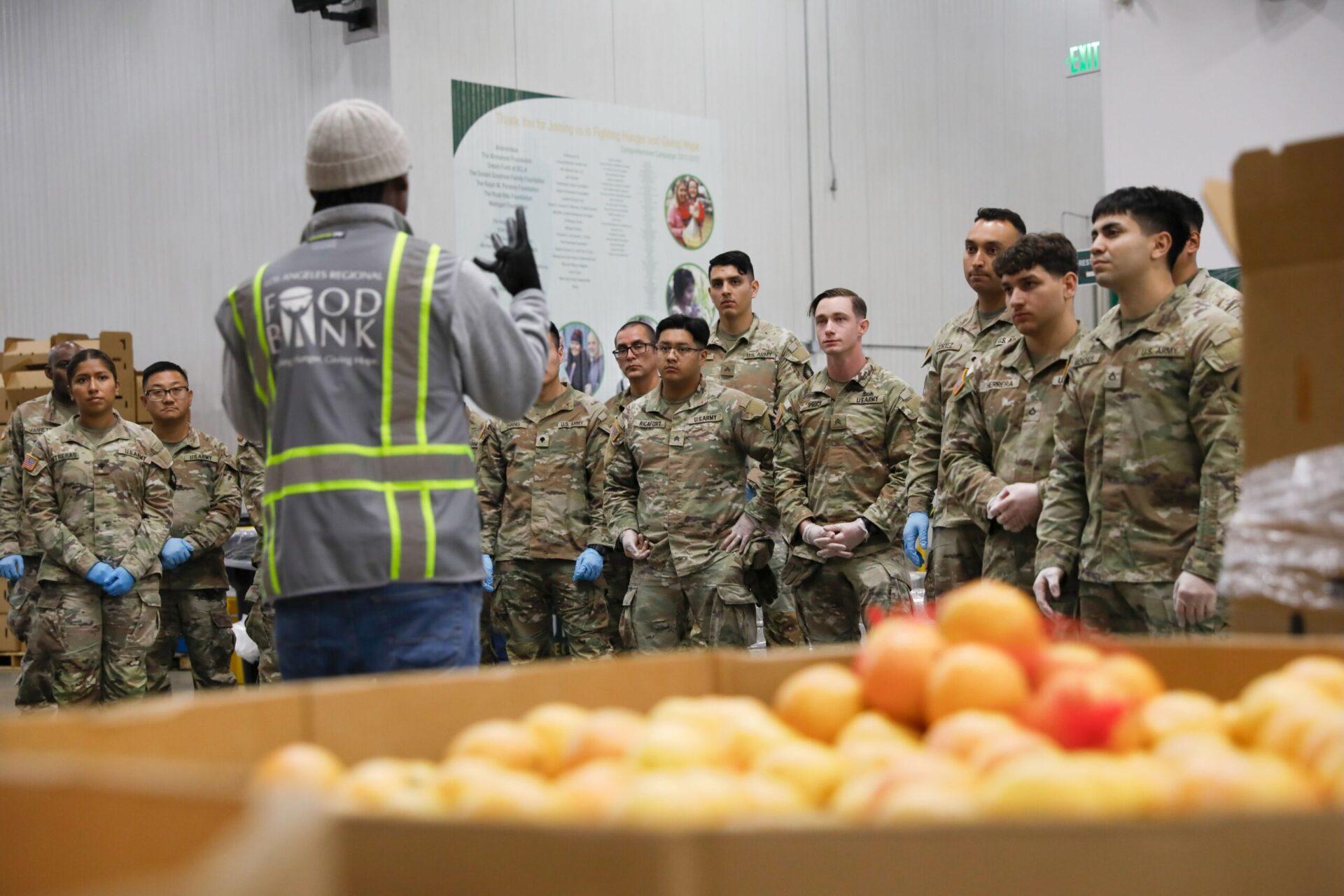 Around 20 members of the California National Guard stand in a warehouse with gloves, listening to an employee of a food bank wearing a gray and lime colored vest and a white beanie.
