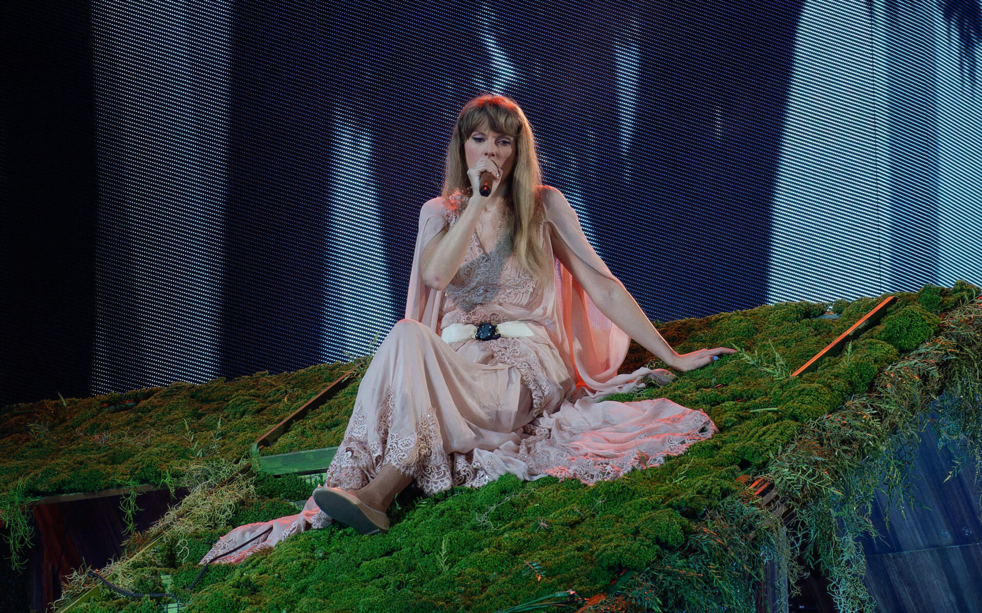 Taylor Swift sings at a concert venue in a pink dress. She sits on a sloping grass lawn.