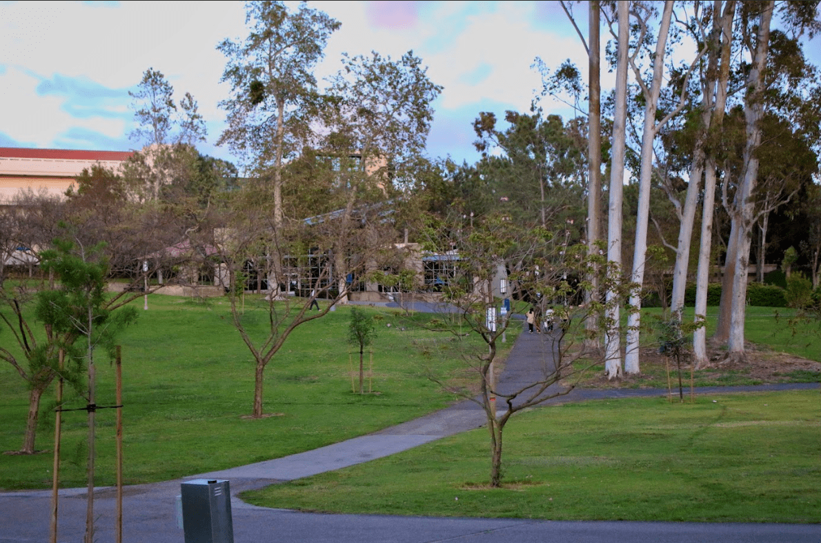 Trees of UC Irvine’s Aldrich Park are shown on a partly cloudy day, students are seen walking along the park’s paths.