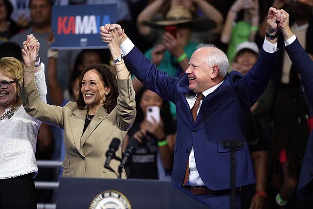 Kamala Harris and Tim Walz hold their hands together in the air at a political rally.