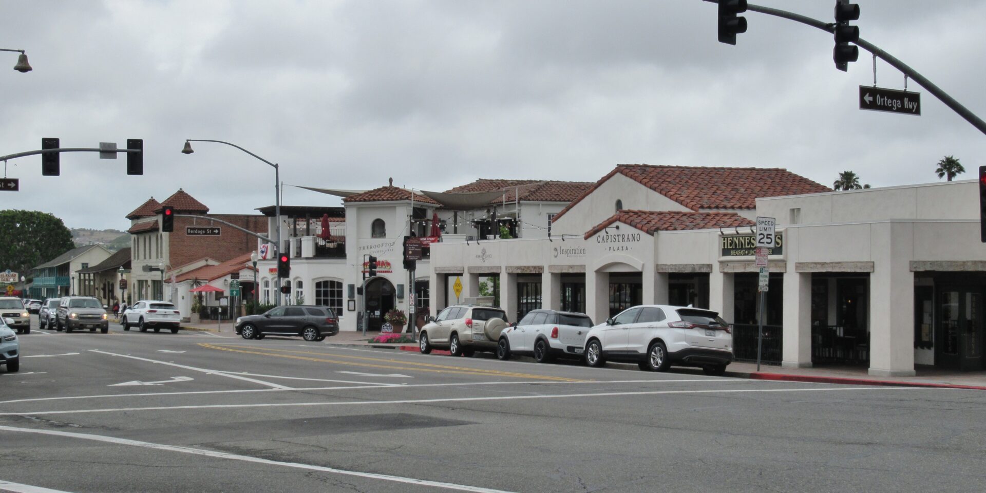 The photo depicts a street in San Juan Capistrano with buildings and cars parked on the right side.