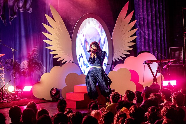 Magdalena Bay lead singer, Mica Tenenbaum, sings at a concert venue in front of a large pair of wings.