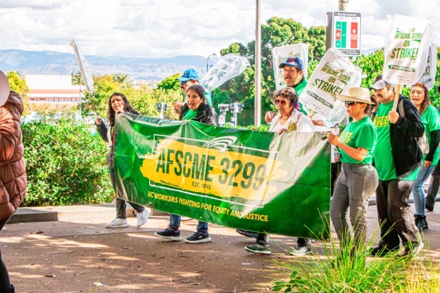 AFSCME workers and protesters are shown walking and holding signs and a large green and yellow banner.