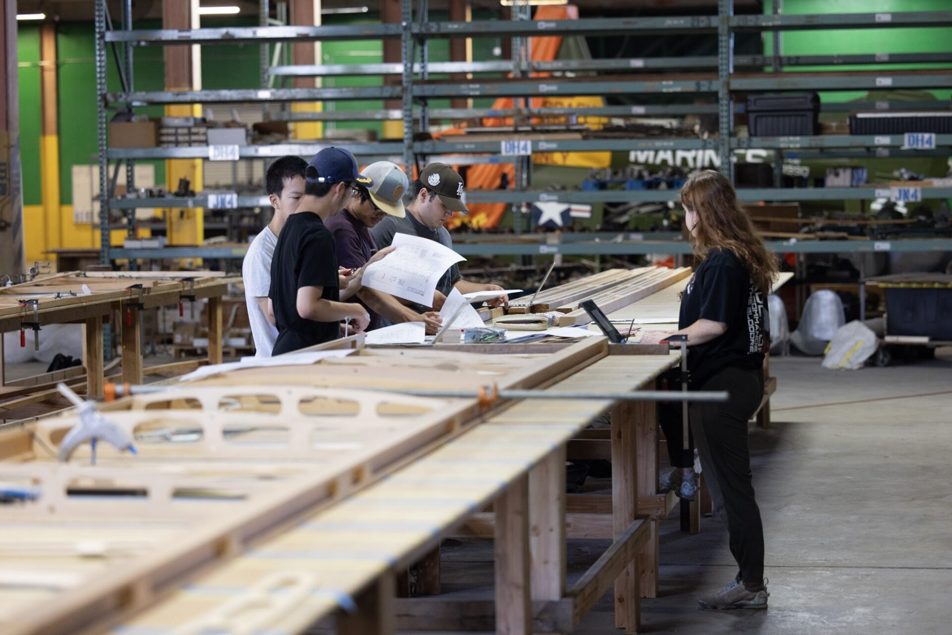 Four UCI students stand on one side of a large wooden table while another student stands on the other side. The students look at blue prints and on their laptops.
