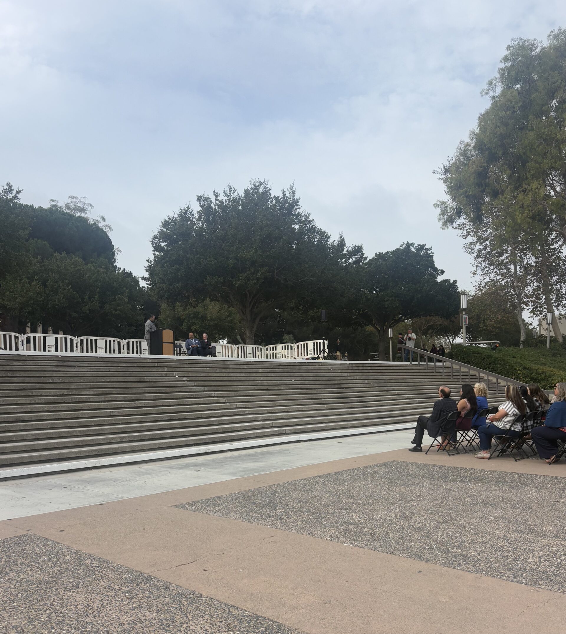 At the stairs by the flagpoles at UCI, a speaker is shown at the top of the stairs behind a podium. At the bottom of the stairs are audience members sitting down.