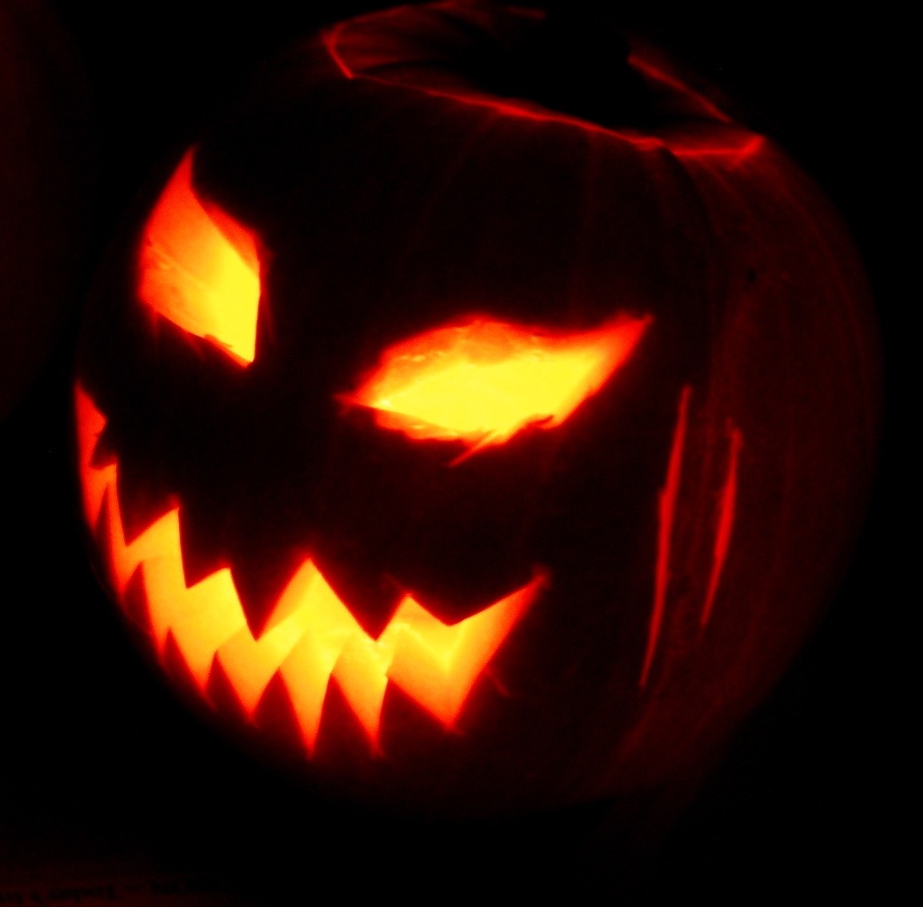 A darkly lit image of a jack-o-lantern with sharp teeth and narrow eyes are shown. The pumpkin looks dark as the eyes and mouth of the jack-o-lantern are the only light source in the image.