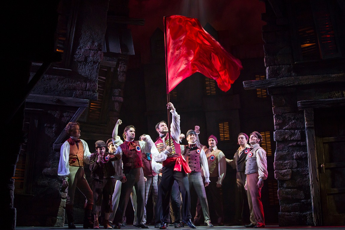 Castmembers of Les Miserables are shown looking up at a flowing red flag being held up by a man at center stage.