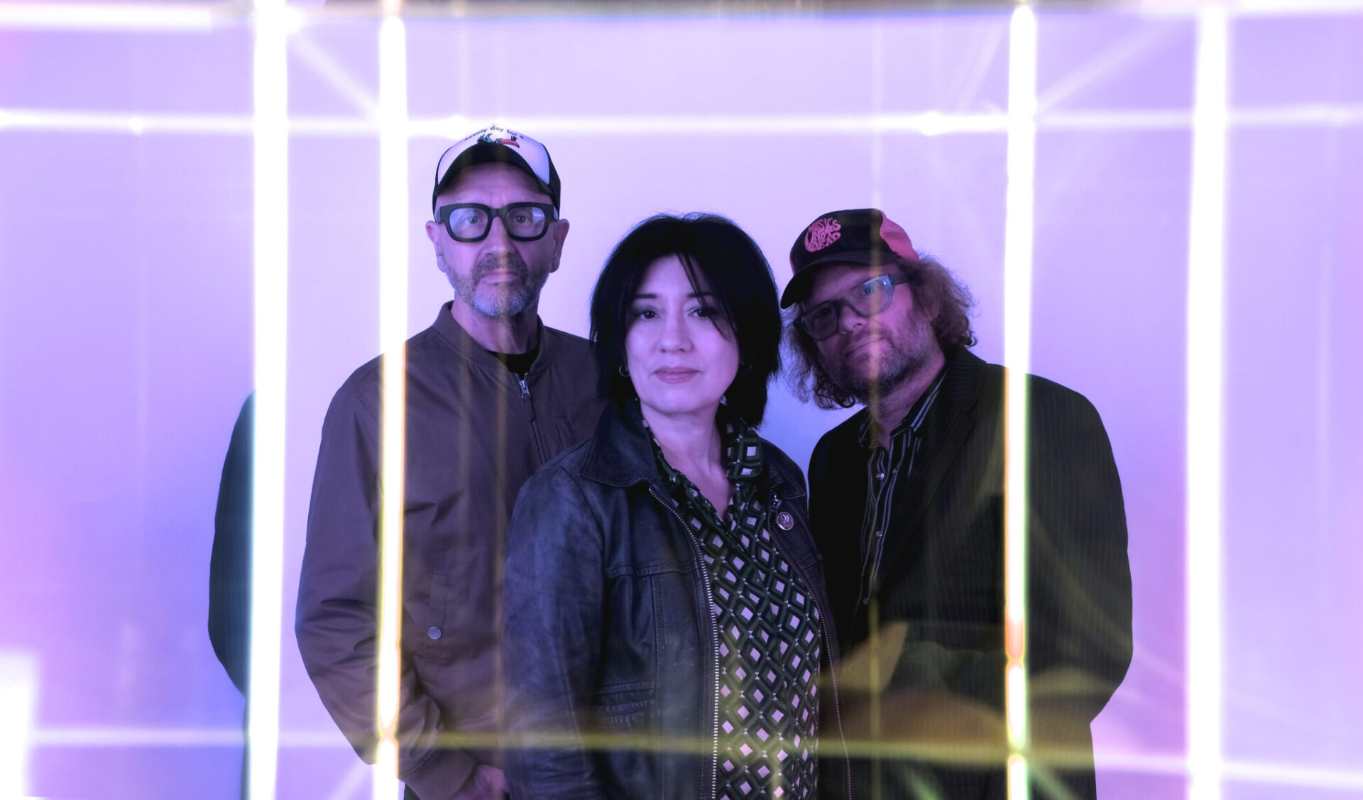 The Miki Berenyi Trio stand in the center of a white room illuminated with pink fluorescent lights.
