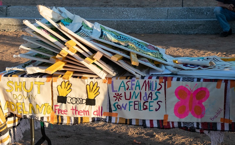 A small table with colorful, neon posters is placed outside the Adelanto ICE processing center, with signs lying on top. The posters say, “Shut down Adelanto” and “Free them all” with a painting of hands in handcuffs.