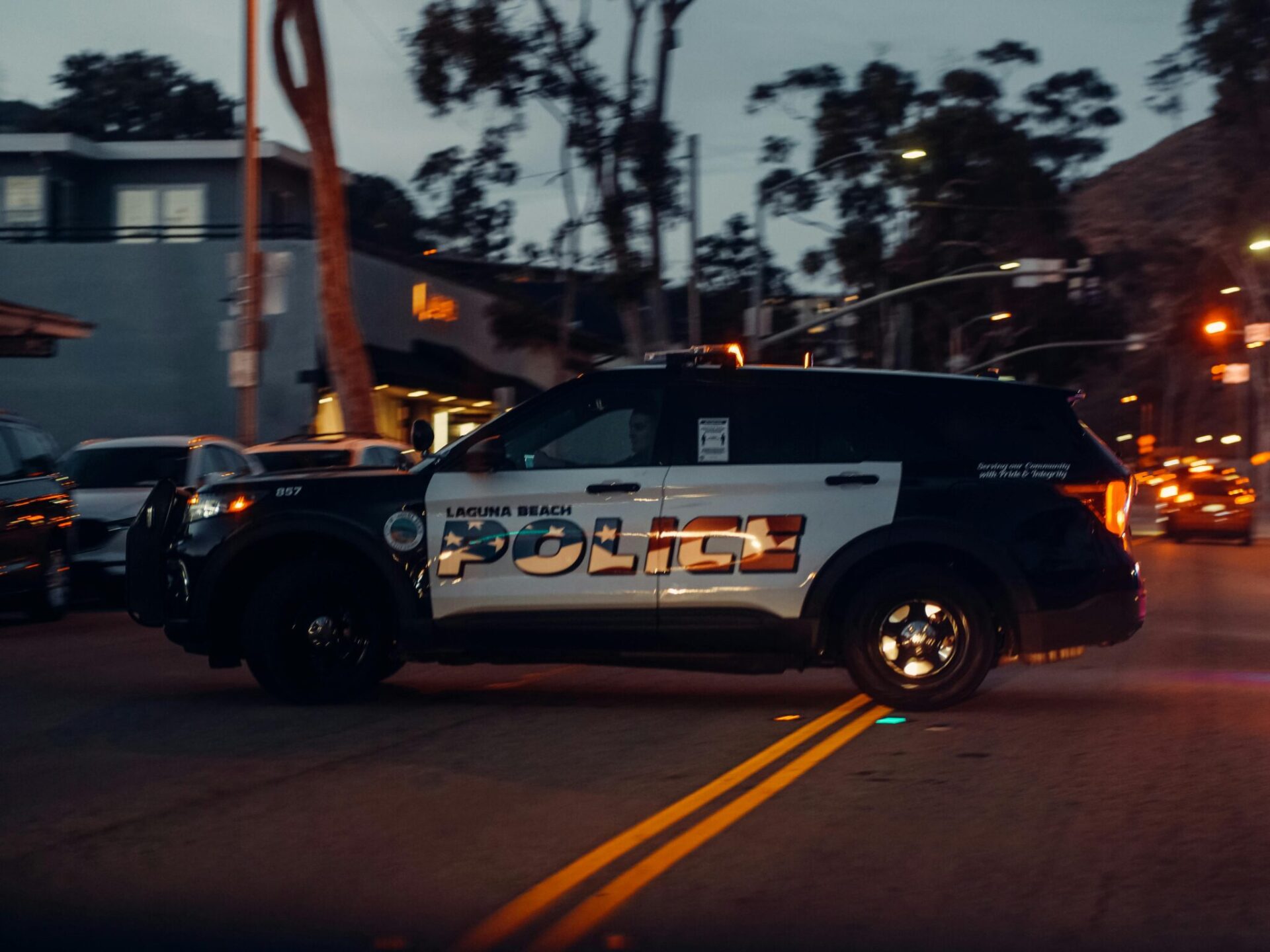A Laguna Beach police car is shown making a U-turn in the middle of a main street in front of a four-way intersection with it’s headlights on, on a late evening.