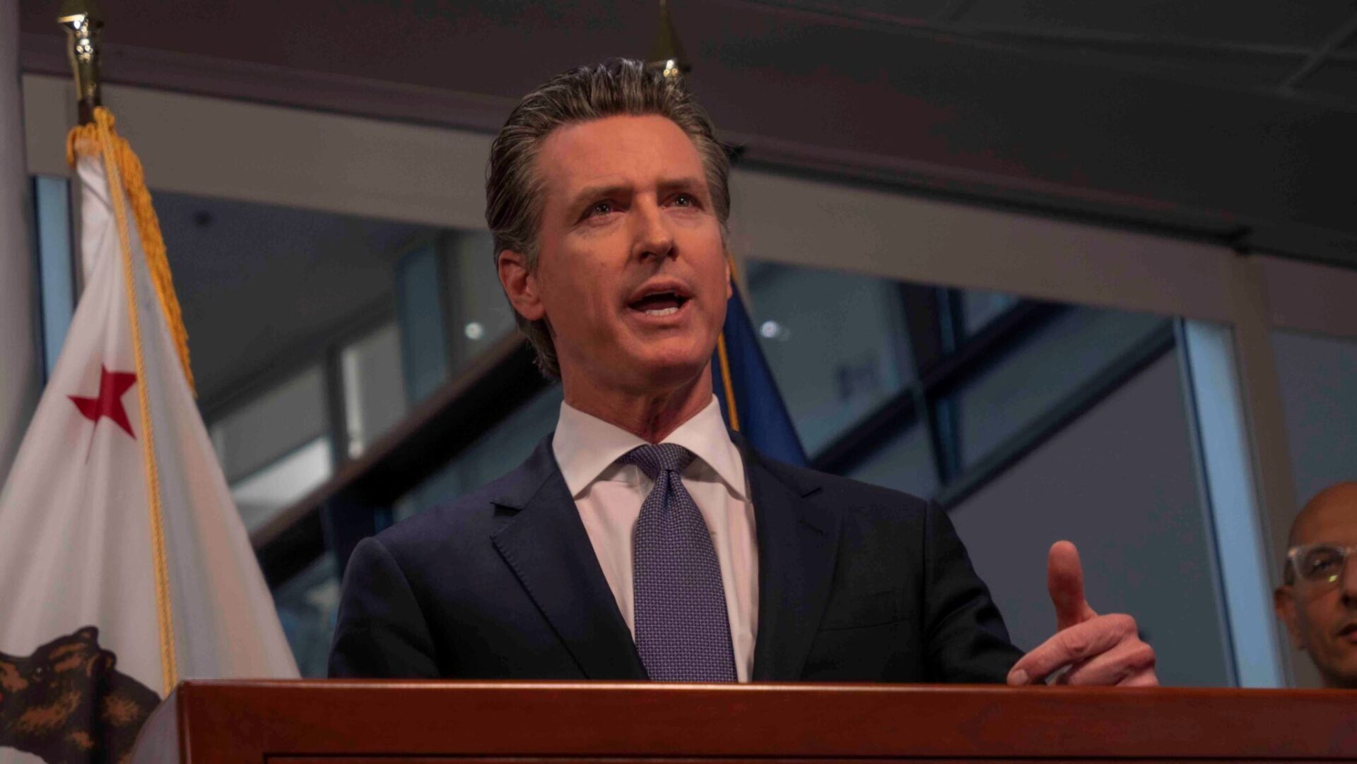 Governor Gavin Newsom stands in front of podium speaking in a dark blue suit and tie, with the flag of California in the background.