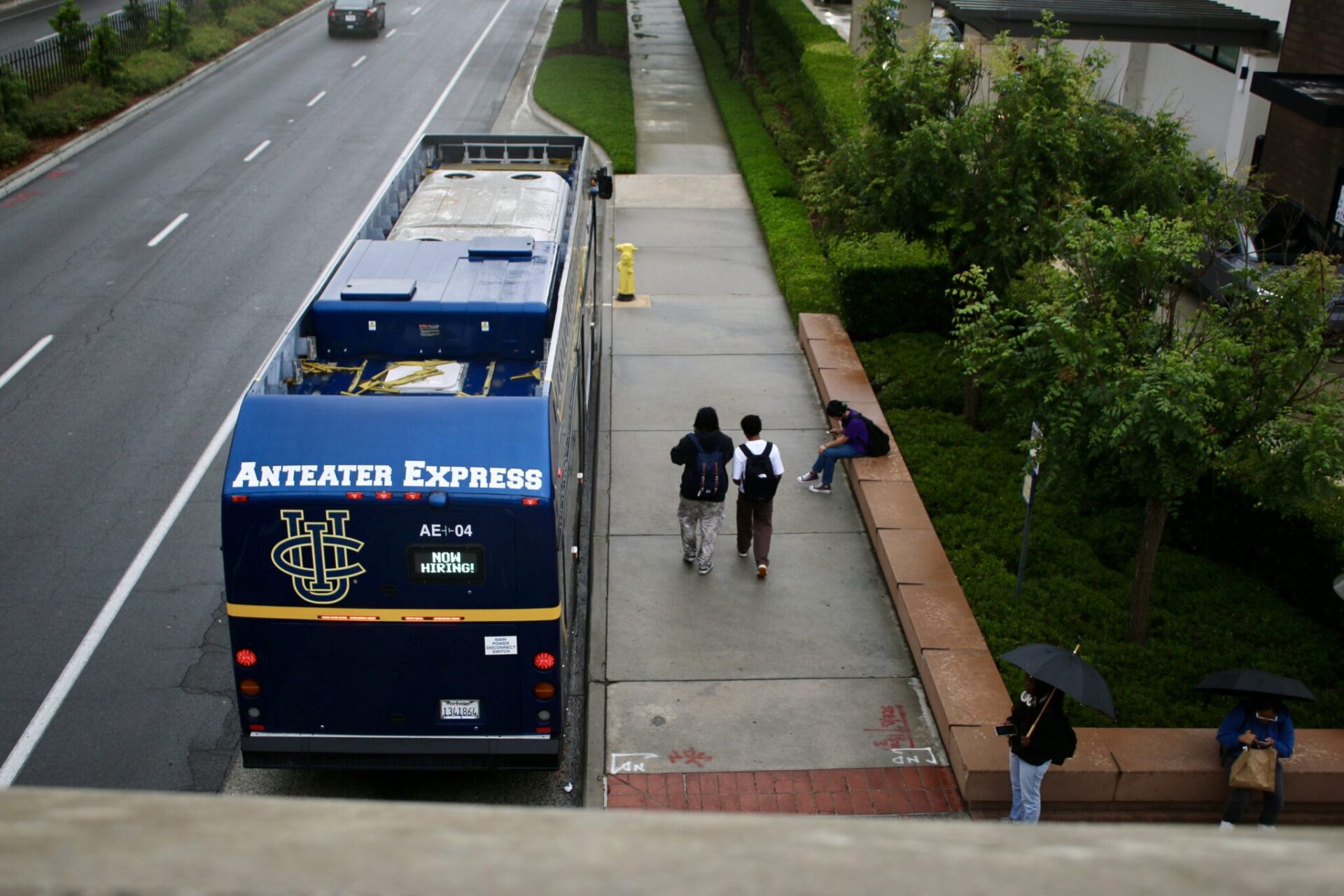 A bird’s eye view of the Anteater Express bus at the University Town Center at UCI is shown parked at the bus stop as students are walking along the side of it, a student is seen with an umbrella.
