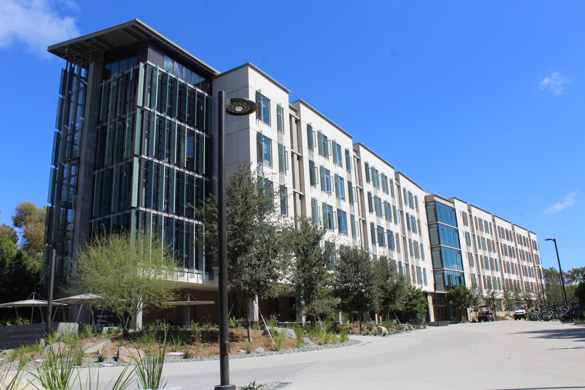 The exterior of Oso Tower at UC Irvine is shown. The building is white with multiple glass windows surrounded by trees and shrubbery.