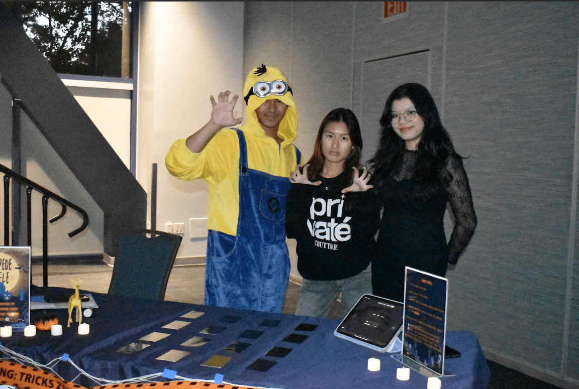 A photo of three UCI students in Halloween costumes stand in front of a display table.