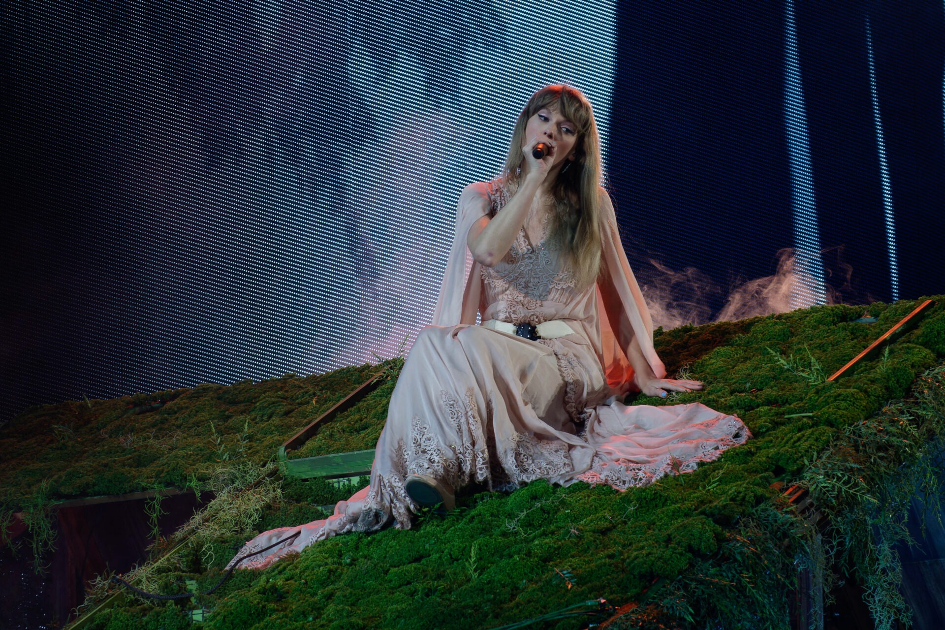 Pop singer Taylor Swift sings on a slanted moss platform wearing a light pink and lace dress. Photo was taken at the Arlington, Texas Eras Tour concert.