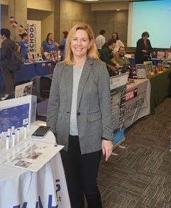 Maryse Mijalski is shown in a grey blazer standing by a round table at a UCI event.