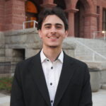Ennes Kahf smiles in front of grey stairs leading to a brown building.