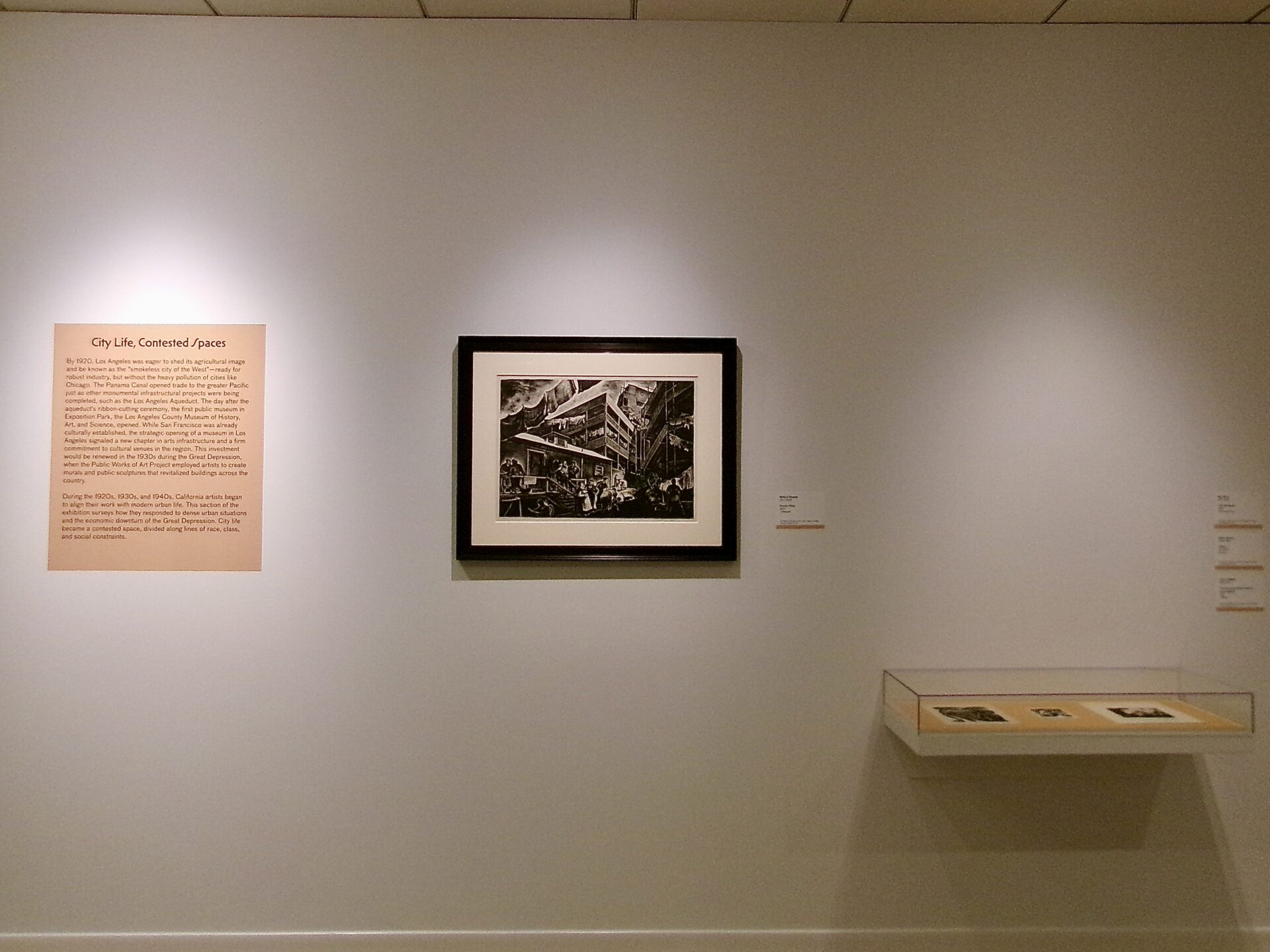 A museum gallery wall displaying a large black-and-white print alongside smaller framed artworks and an informational text panel.