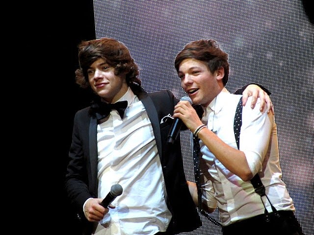 Harry Styles wears a white dress shirt with a black tuxedo jacket and black bow tie holding a microphone with his arm around Louis Tomlinson. Louis wears a white dress shirt buttoned to the top with black-and-white polka-dot suspenders, one arm around Harry’s back and the other holding a microphone to his mouth as he sings.