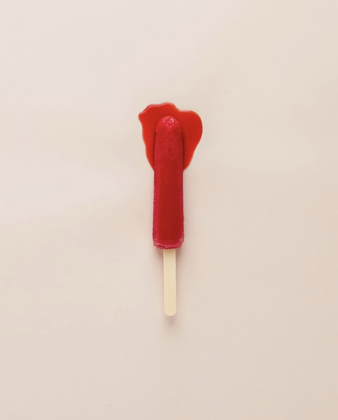 A red popsicle slightly melted at the top and on a wooden stick, centered against a pale pink background.