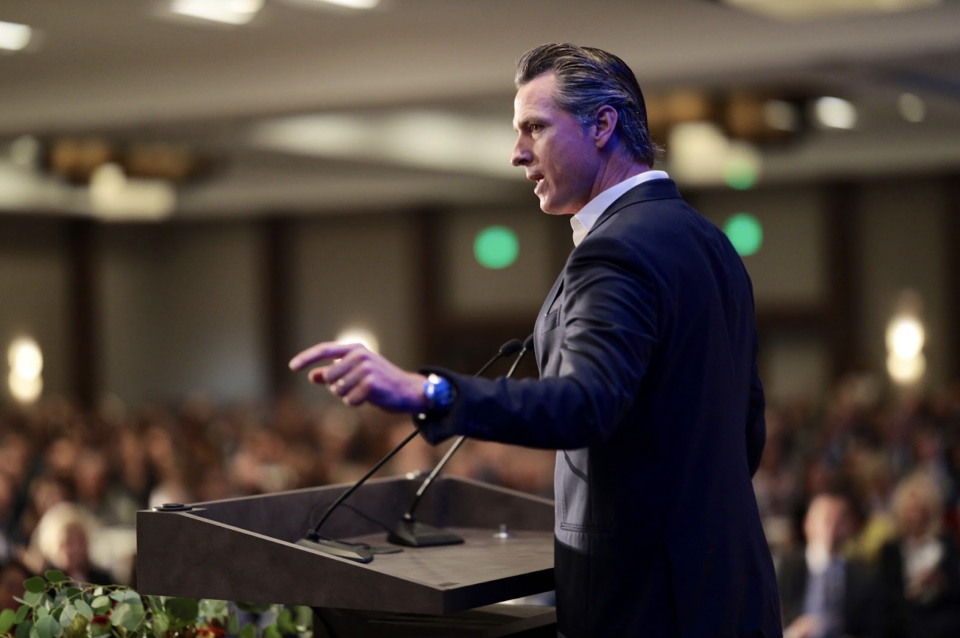 A waist-up photo of Gavin Newsom standing at an indoor podium with his left shoulder facing the camera. He speaks into two chest-level microphones and points slightly to his left side. A crowd sits to his front and sides, out of focus.