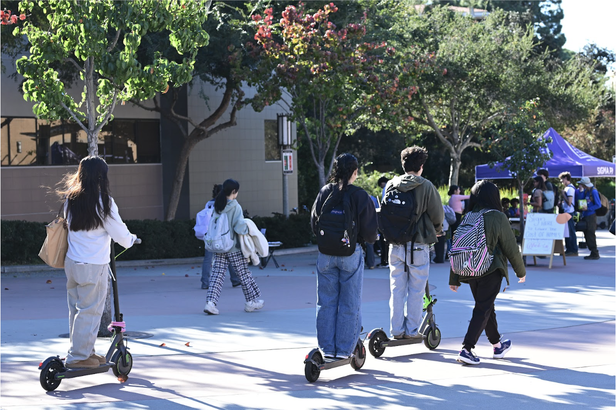 Three students on electric scooters and two students walking on Ring Road head toward the same direction.
