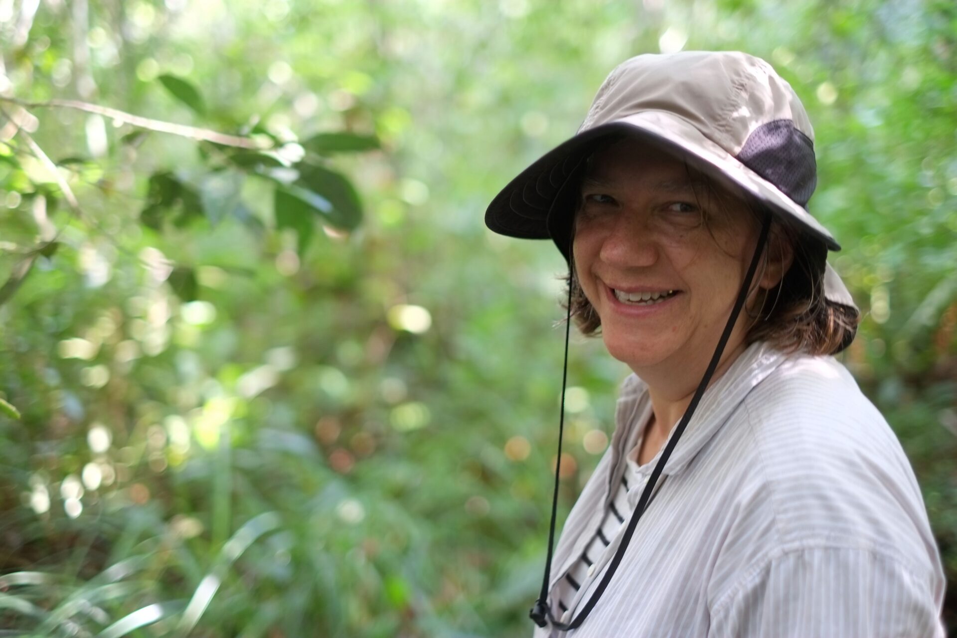 Susan Trumbore smiling in a tan bucket hat and light-colored shirt, standing in a lush tropical forest setting with green foliage in the background.