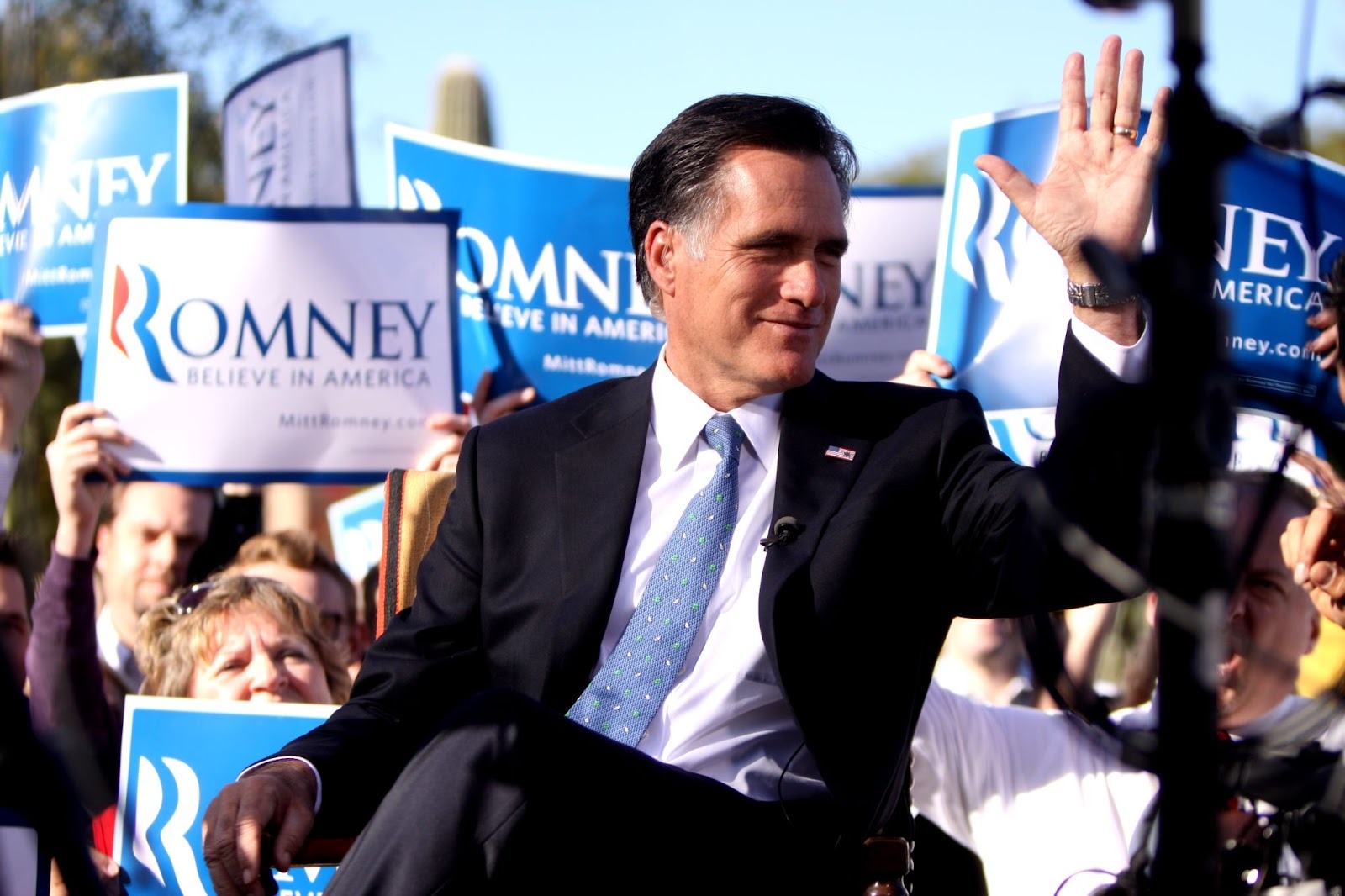 Mitt Romney is in a dark suit and light blue tie. He sits elevated above a crowd at an outdoor political rally, raising his right hand in a wave. He is turned slightly to his left and smiling as supporters stand closely behind him holding blue campaign signs that read “Romney” and “Believe in America.” The crowd fills the background, with signs raised at varying heights, suggesting an energetic campaign event in a sunny, desert setting.
