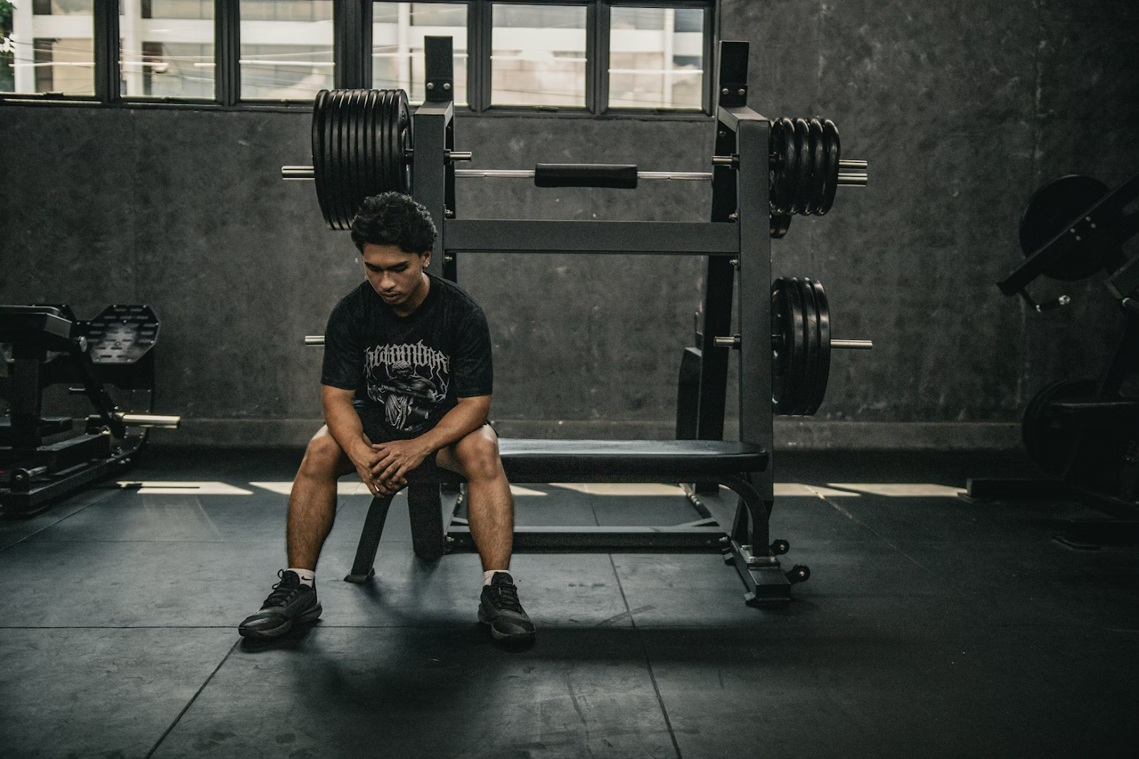 man in athletic attire sits on the edge of a plate-loaded squat rack with his elbows on his knees and his hands crossed, as he looks down toward the floor.
