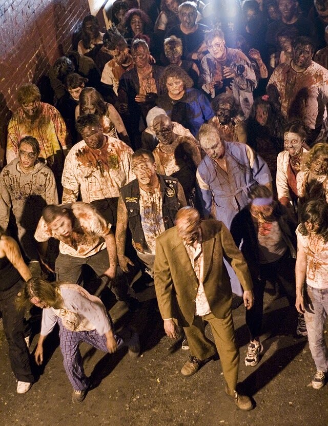 A mob of extras dressed as zombies stagger forward. Some wear white facial make, while most are blood stained. They vary in clothing style.