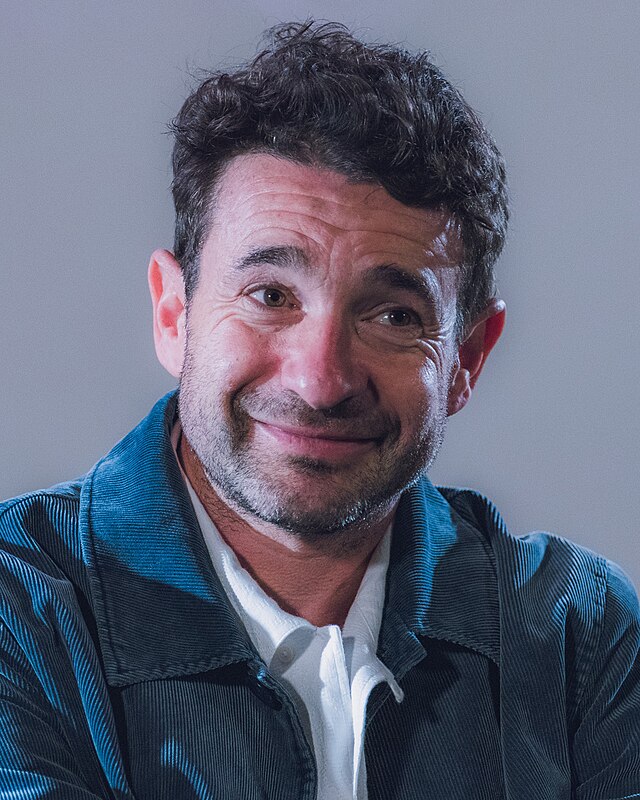 A photo of Bart Layton, the director of Crime 101, at a press conference. He is smiling beyond the camera, wearing a white shirt with a blue button-up.
