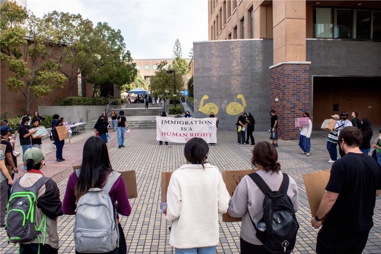 College-aged students holding signs gather in a circle around a large banner that reads “IMMIGRATION IS A HUMAND RIGHT.”