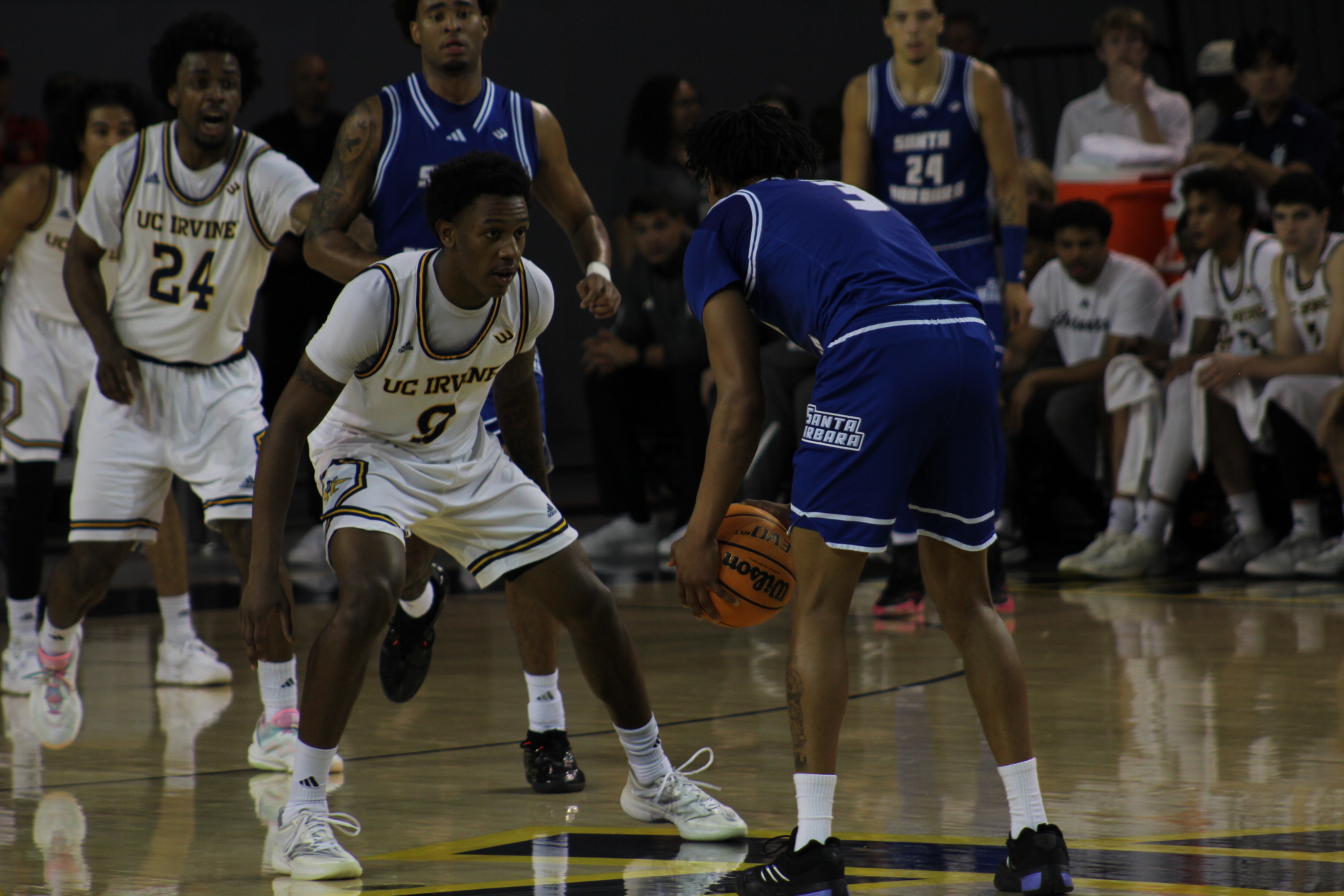 The opposing Santa Barbara player dribbling the ball while UCI's #9, Jovan Jester Jr. blocks his path to the basket and takes a defensive stance.