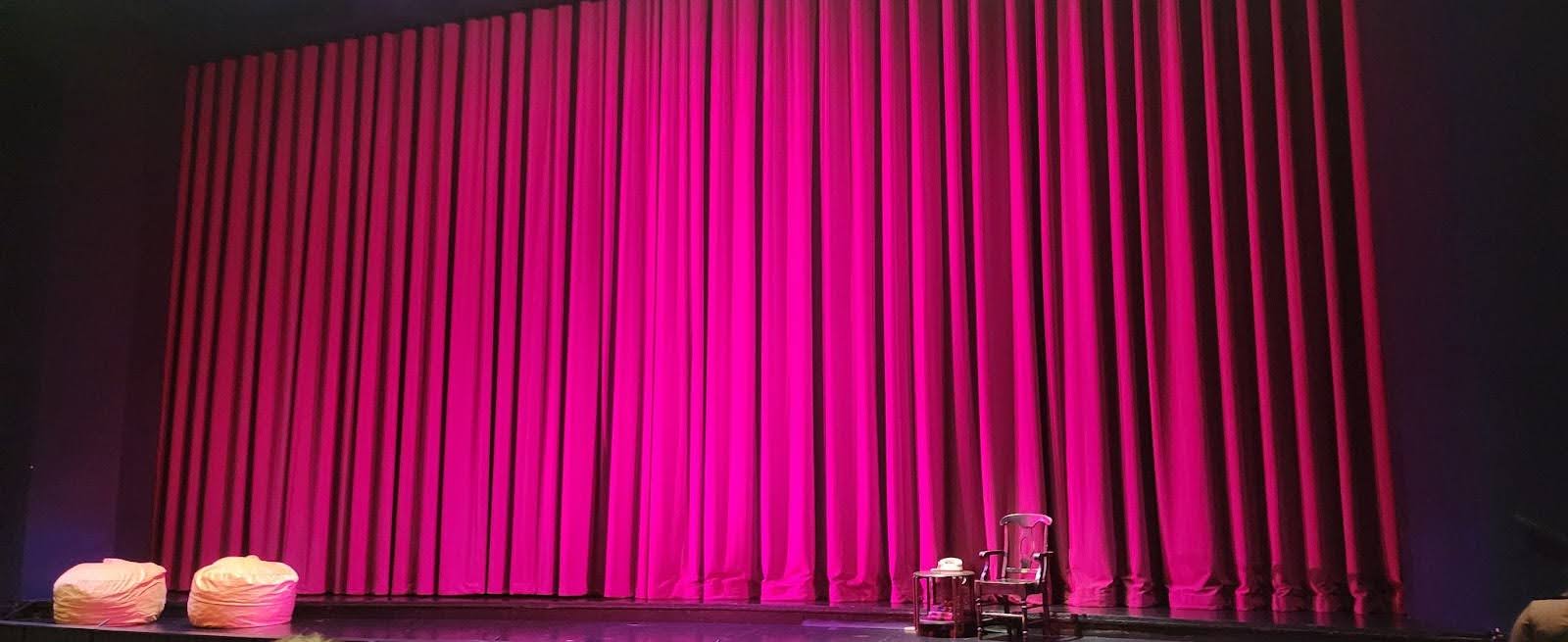A photo of a stage with a lowered red curtain. The center is illuminated by spotlight. A wooden chair and rotary phone also sit onstage.