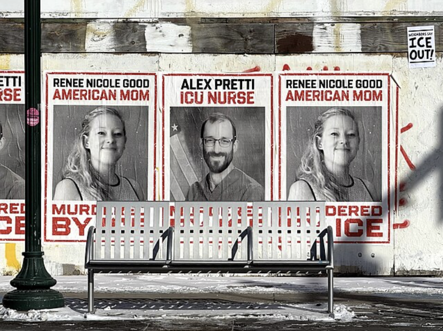 A poster is hung up on a graffitied plywood wall, depicting black and white photos of Alex Pretti and Renee Nicole Good with red text saying “Murdered by ICE.” In the upper righthand corner of the image, a much smaller poster reads “ICE OUT!” A bench and lamppost partially block the poster from view.