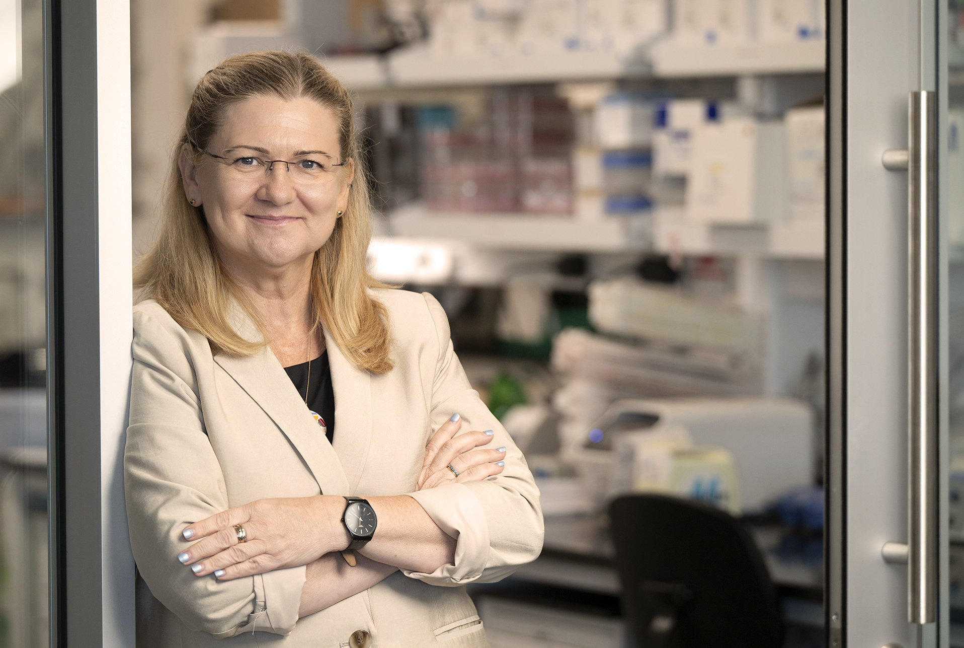 A professional headshot of a woman with shoulder-length blonde hair and glasses, wearing a cream blazer over a black top, standing with her arms crossed and smiling slightly. The background shows a blurred laboratory setting with shelves of supplies and equipment.