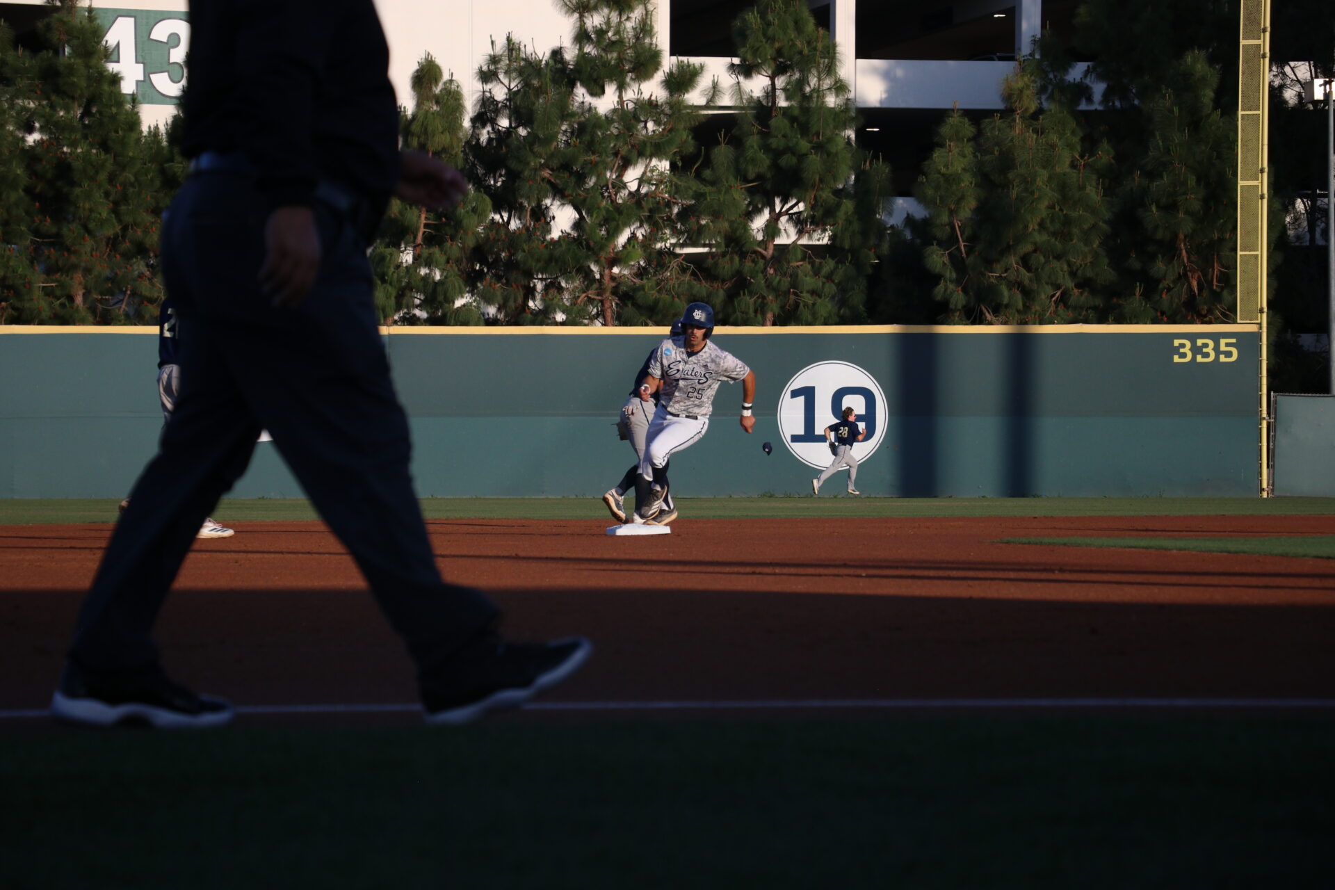 UC Irvine player number twenty five Alonso Reyes speeds down second base as UC Davis player number twenty eight is behind him in the outfield running to catch the ball which causes his hat to fly off his head.