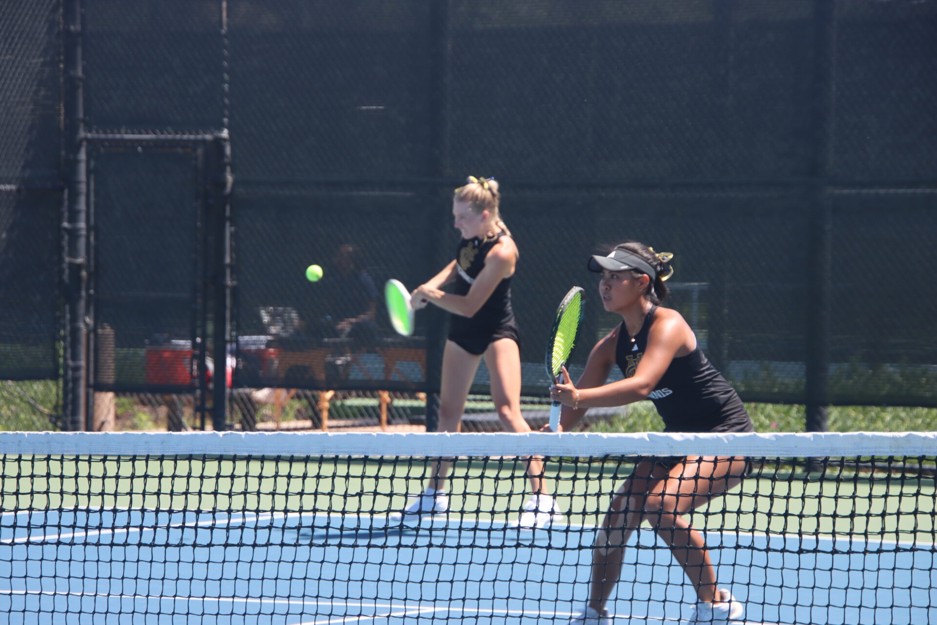 Two UC Irvine women's tennis players stand ready at the net on an outdoor court, holding rackets and facing their opponents during a doubles match against UC Santa Barbara