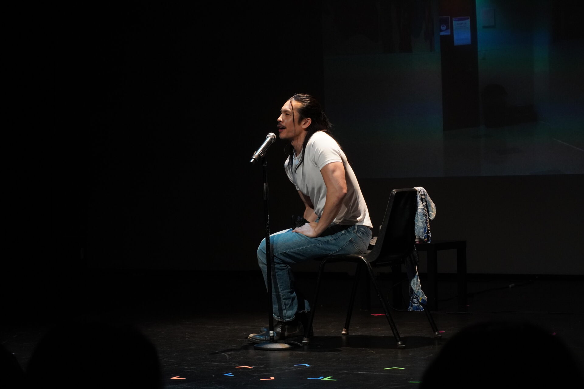 Kanazawa leans towards, from a black plastic chair on stage, a microphone. He is wearing a white tee, blue jeans, and black boots.