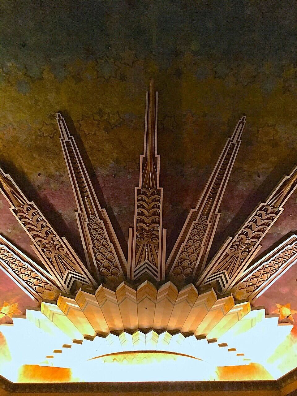 A photo of a sun motif that is part of the Wiltern theater's architecture. This image is illuminated by lights at the bottom and brown, triangular spikes come from the bottom arch.