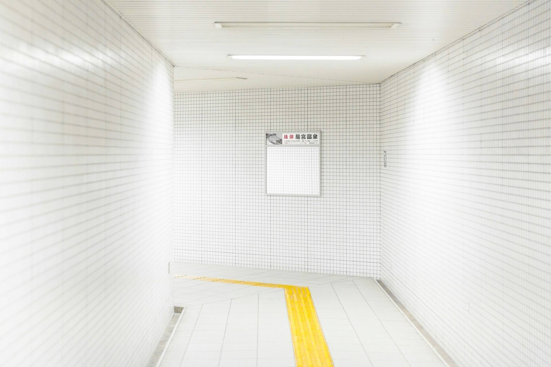 A white, tiled hallway leading to a left turn. There is a yellow line dividing the middle of the hallway and it leads to a white sign up ahead.