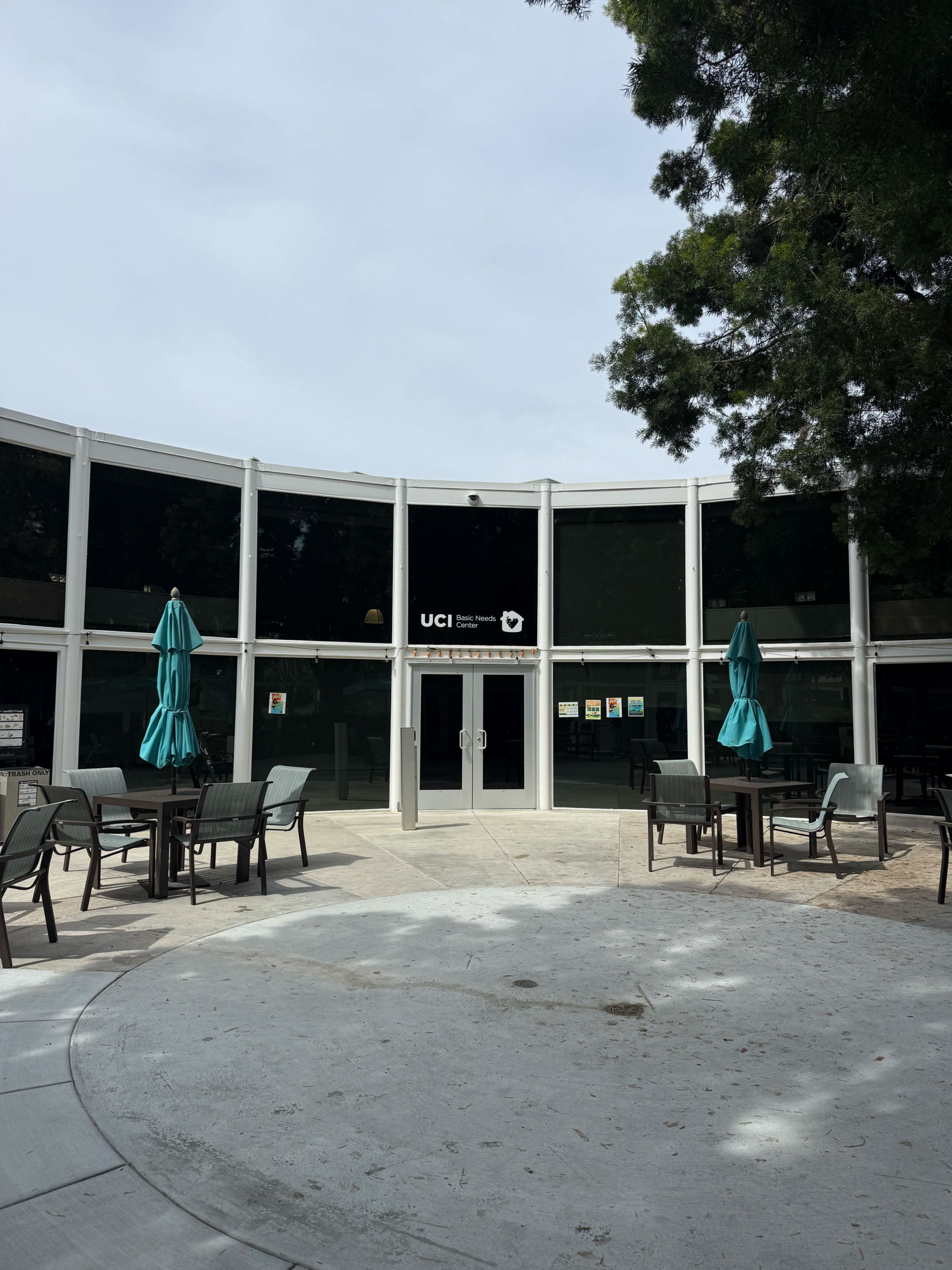 Exterior view of the UCI Basic Needs Center, a curved glass-front building with dark tinted windows. Several outdoor tables and chairs with closed teal umbrellas sit on a concrete patio in front, with trees partially framing the scene under an overcast sky.