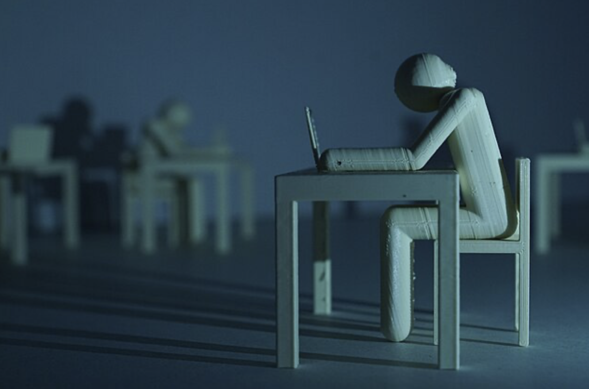 A 3D-printed figure of a person sits on a chair pushed in at a desk, head bent and shoulders slumped over a computer in front of them. The desk casts long shadows to the left and over the figure’s head. In the background, a blurry, blue-and-grey-tinted identical figure can be seen