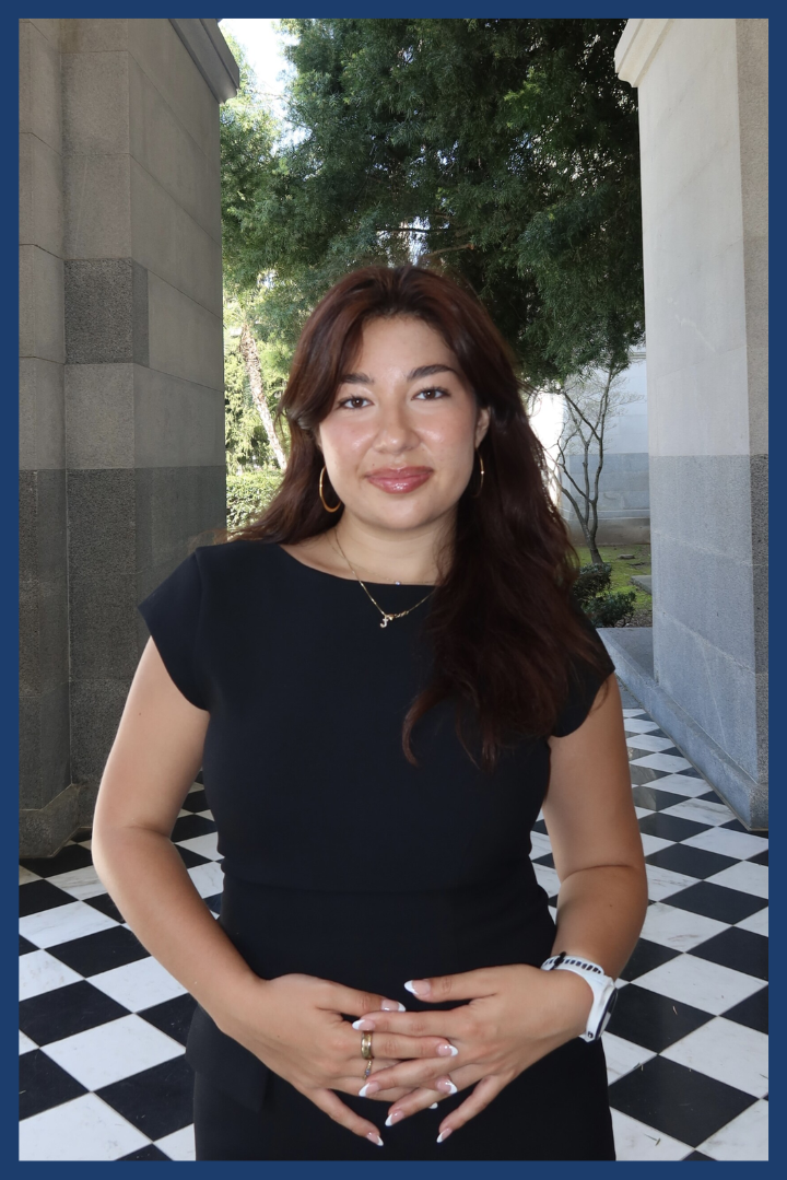 A photo of ASUCI External Vice President candidate Jayla Kamilla Pete is shown. Pete wears a black shirt standing in a hallway with checkered floors.