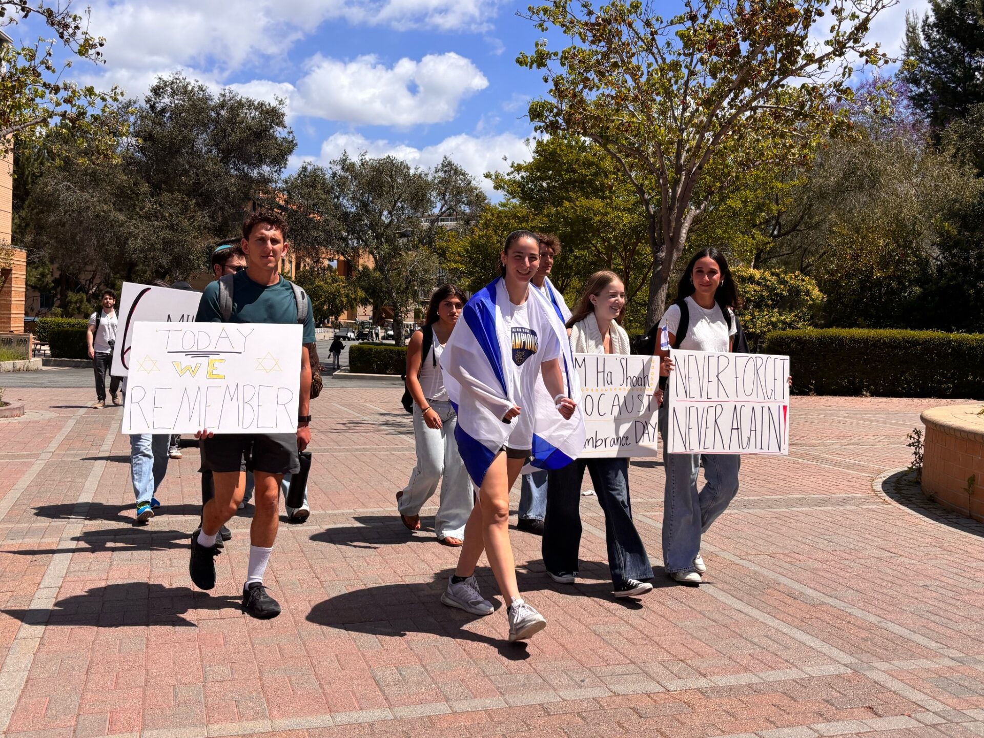 UCI students walk on campus on brick floors with signs reading “TODAY WE REMEMBER” and “NEVER FORGET NEVER AGAIN!” One student wears an Israeli flag over her shoulders while others wear white for the event.