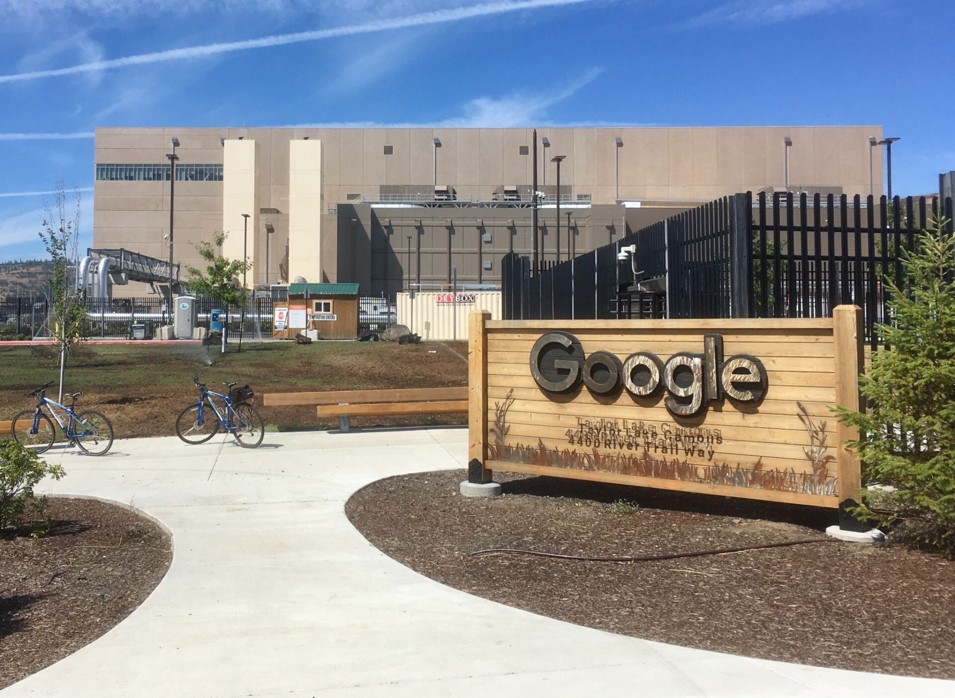 The Google logo stands on a wooden sign outside a large data center building, with security fencing, pipes and equipment visible, and two bicycles parked along a sidewalk in the foreground under a clear sky.