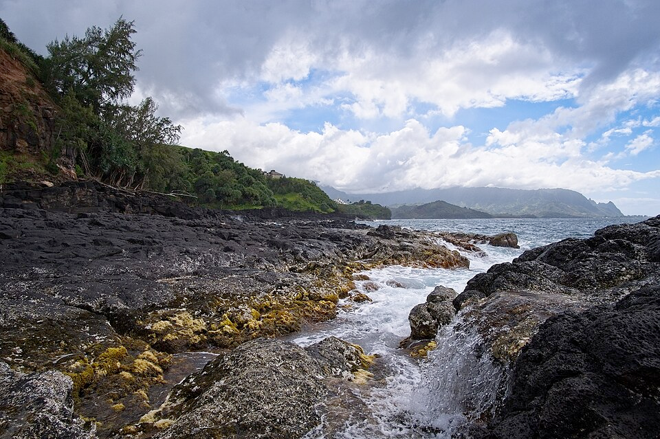 A small stream of water leads out into the ocean around the island of Kauai, surrounded by black rocks with foliage and mountains in the background.