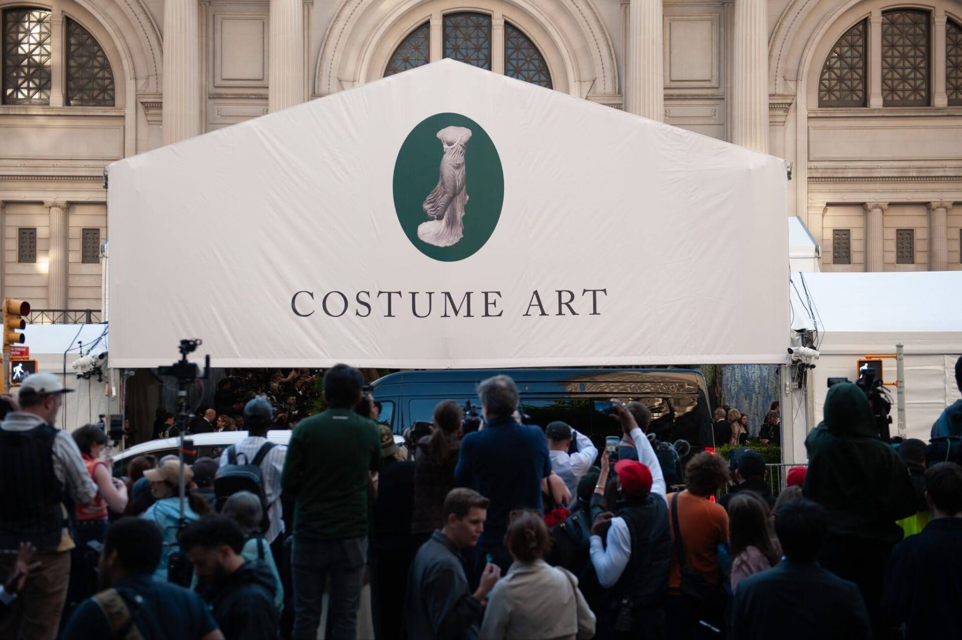 A crowd convenes at the Metropolitan Museum of Art for the 2026 Met Gala. On a canvas cover above the entrance is written 